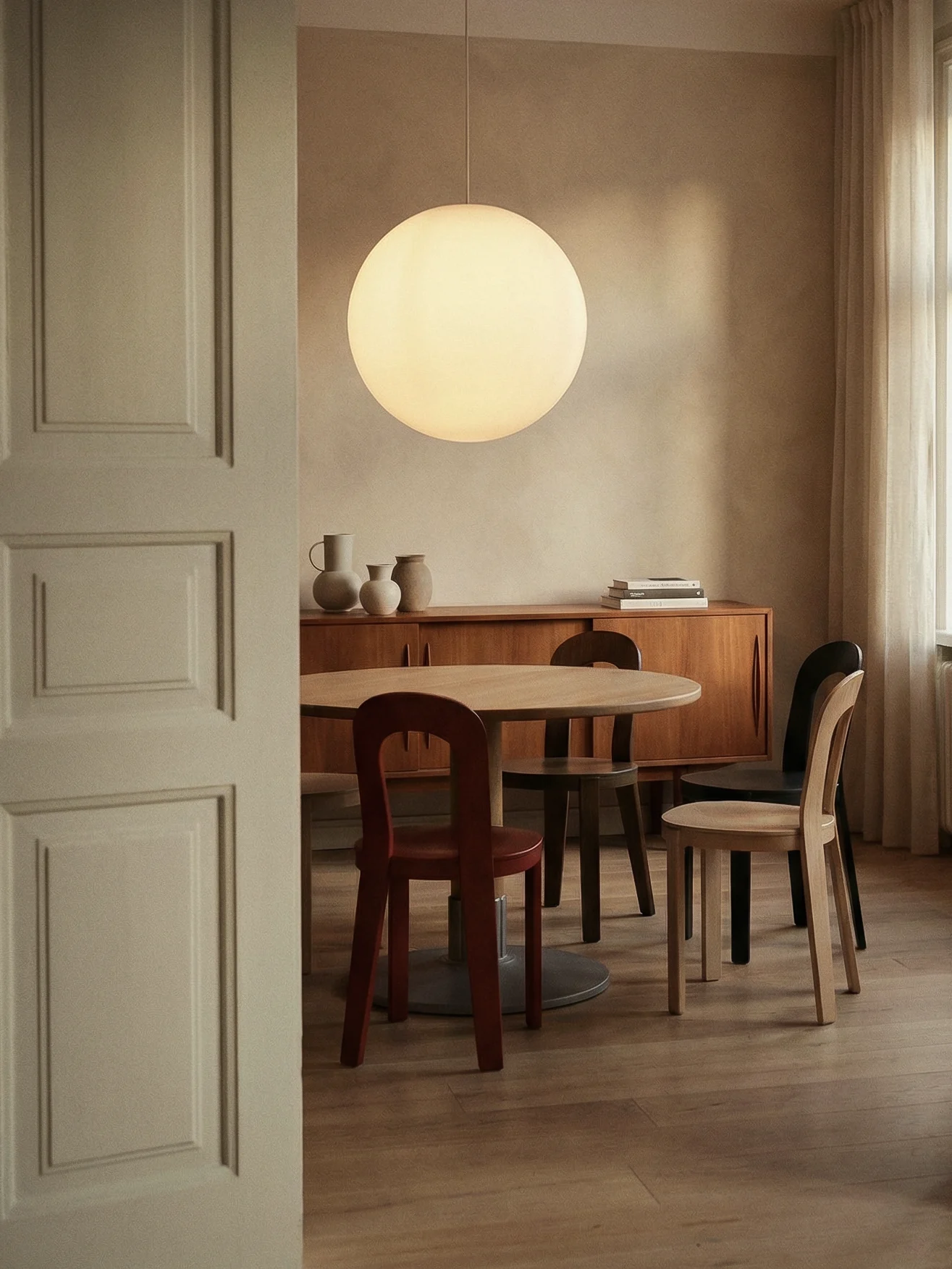 Uma sala de jantar moderna com mesa redonda de madeira, cadeiras variadas, um grande candeeiro suspenso esférico e uma credência com vasos decorativos.