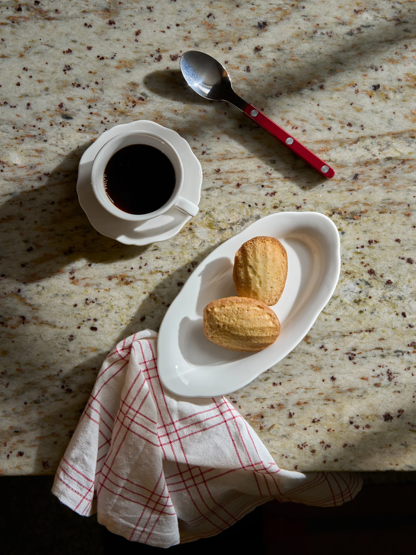 Vista aérea: café preto em xícara branca, duas madeleines em prato branco, colher e guardanapo listrado sobre mármore.