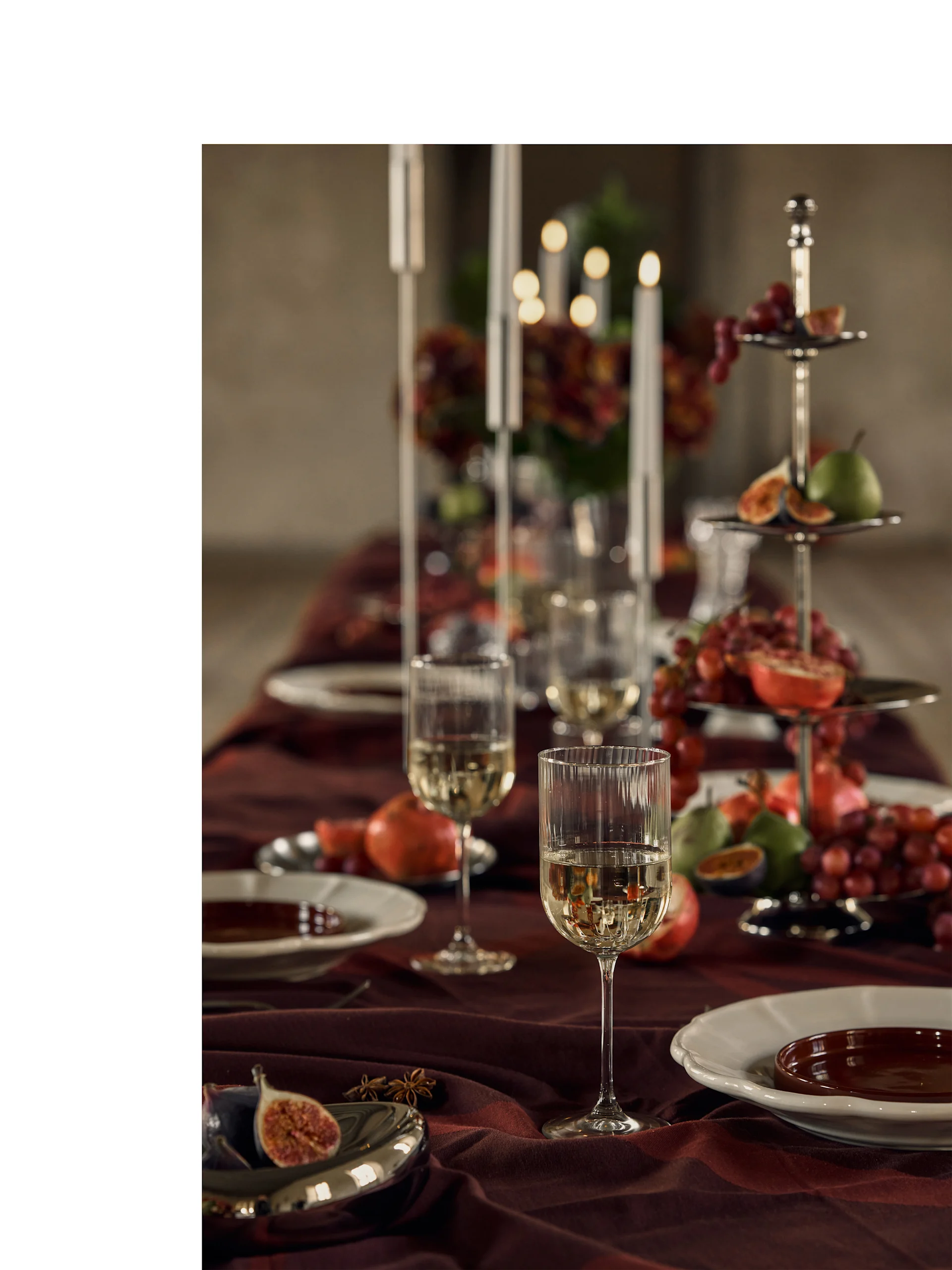 Mesa festiva posta com copos de vinho branco, pratos e um suporte de vários andares cheio de frutas coloridas e luz de velas.