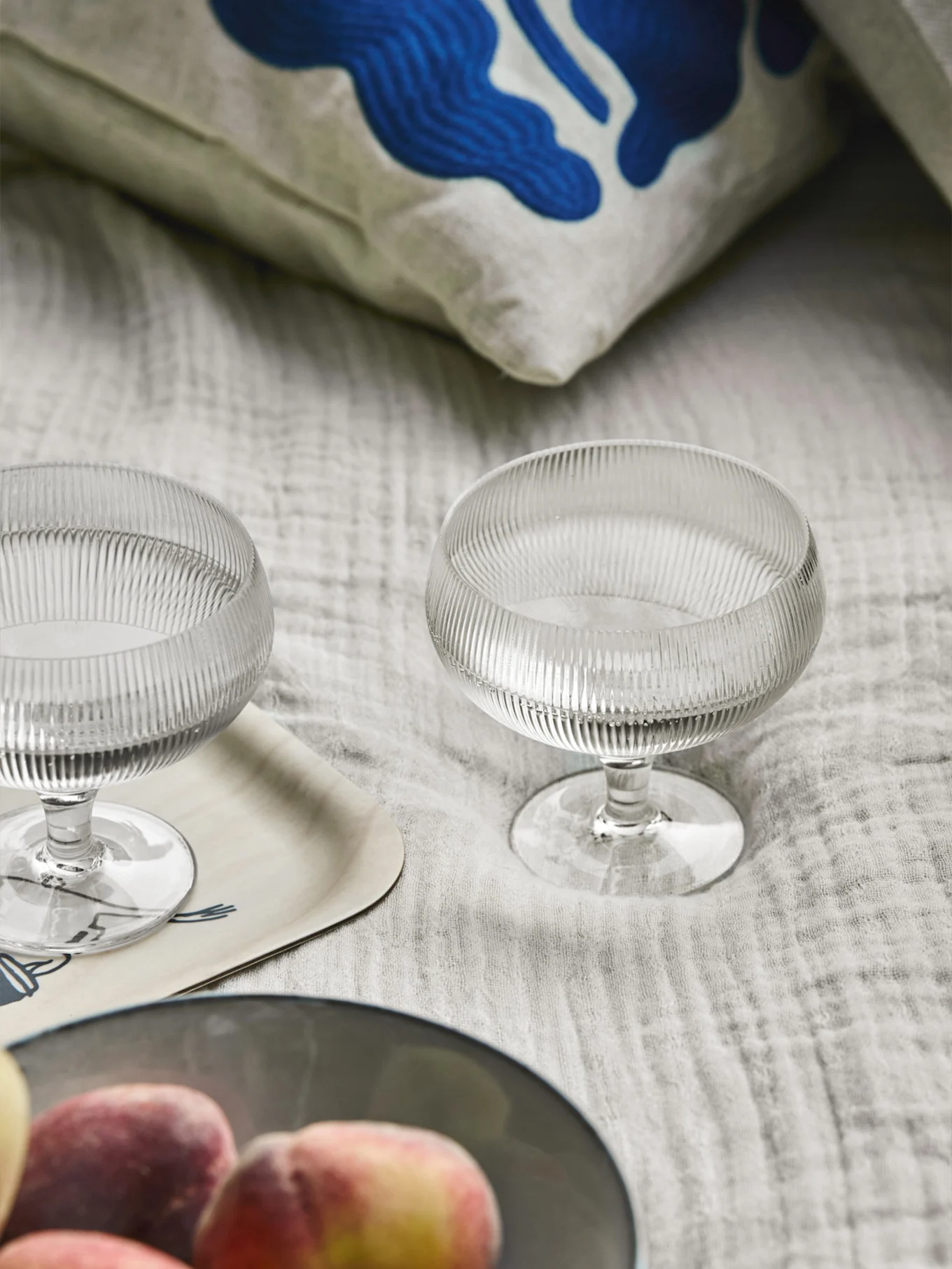 Duas taças de sobremesa de vidro canelado, uma almofada com arte abstrata azul, um tabuleiro de madeira e pêssegos sobre um tecido claro.