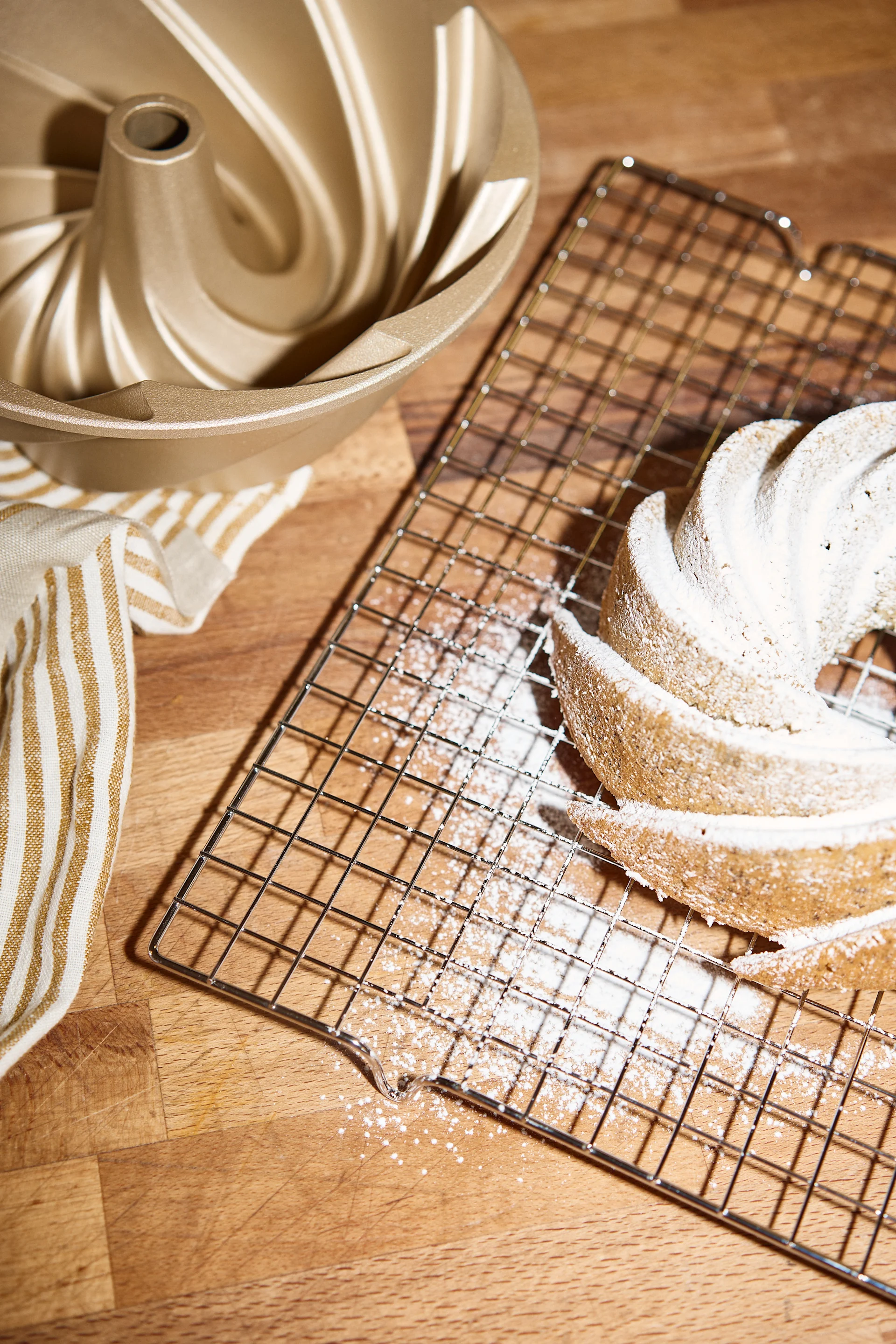 Uma forma bundt dourada, uma toalha listrada e um bolo bundt polvilhado com açúcar em pó sobre uma grade de resfriamento.