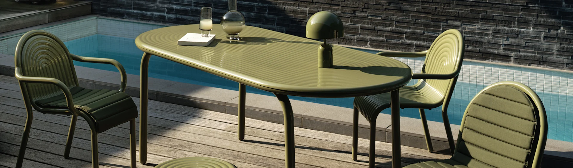 Mesa e cadeiras de jantar verdes caneladas ao ar livre num deque de madeira junto a uma piscina. Um candeeiro, garrafa, copo e livro estão na mesa.