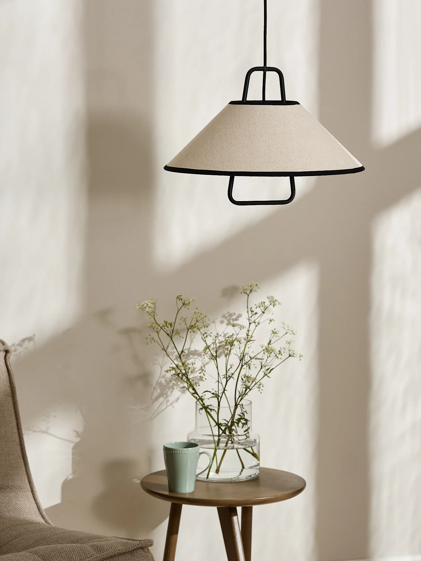 Um candeeiro pendente cónico creme com acabamento preto paira sobre uma mesa lateral de madeira com um vaso de flores brancas e uma caneca verde clara.
