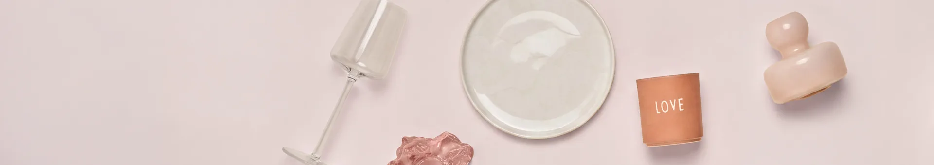 Vista de cima de vários objetos decorativos e de mesa sobre uma superfície rosa claro. Da esquerda para a direita: um copo transparente inclinado, um prato creme, um castiçal de terracota "LOVE" e uma escultura abstrata rosa. Um objeto ondulado rosa menor está sob o copo.