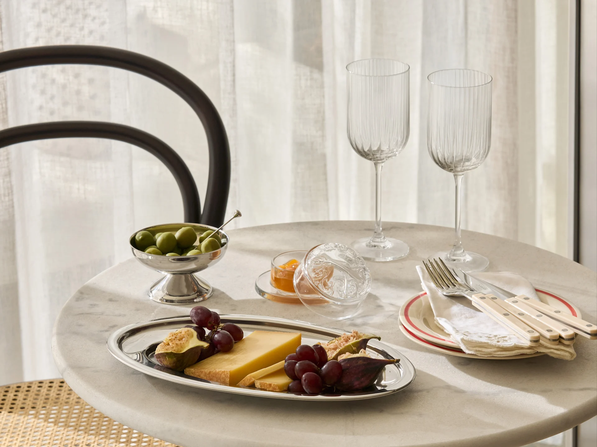 Uma mesa de mármore posta com uma travessa de prata com queijo, uvas, figos, uma tigela de azeitonas, geleia, dois copos estriados e talheres em pratos.