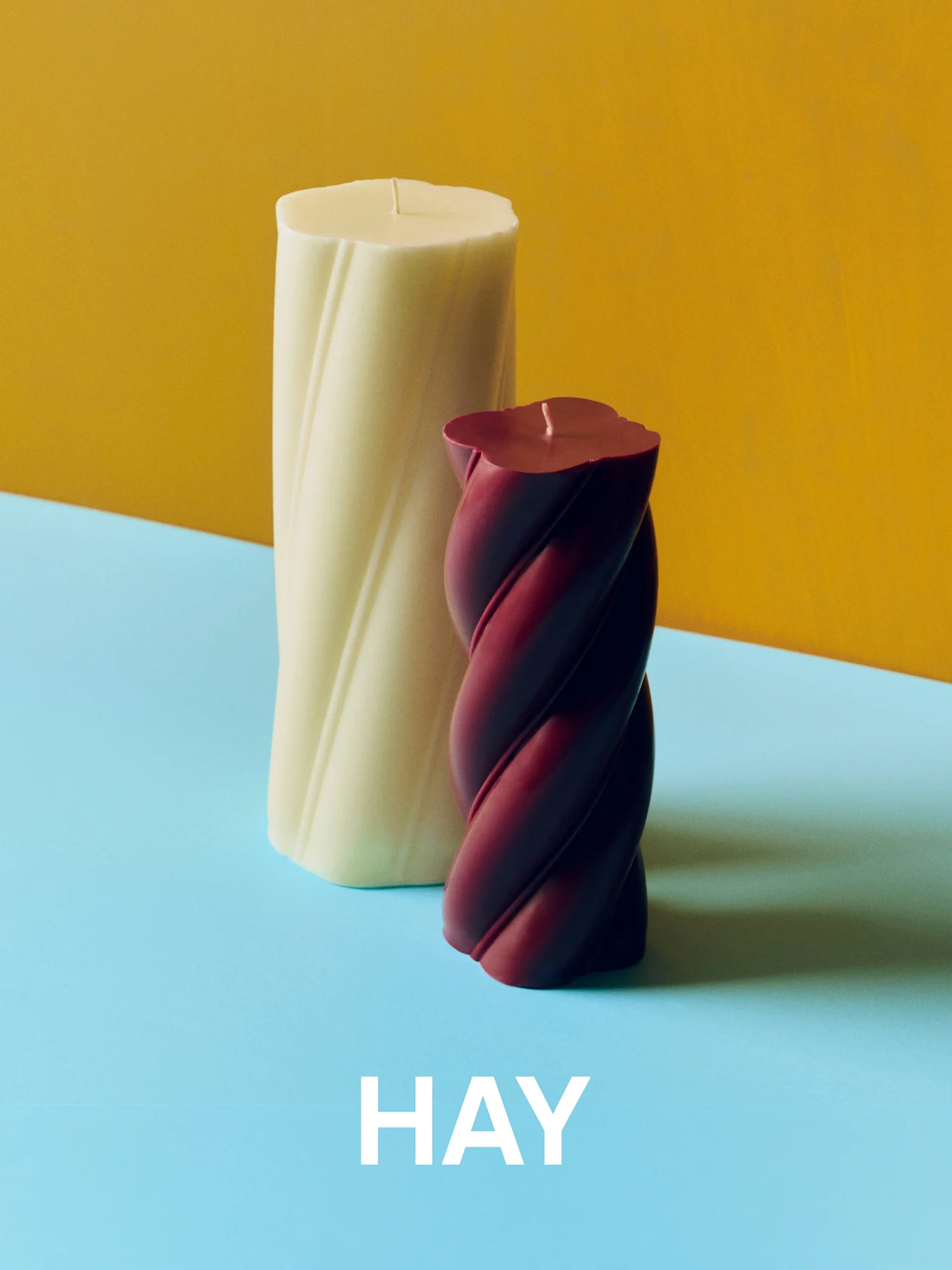 Velas torcidas creme e bordô sobre um fundo amarelo e azul com o texto 'HAY'.