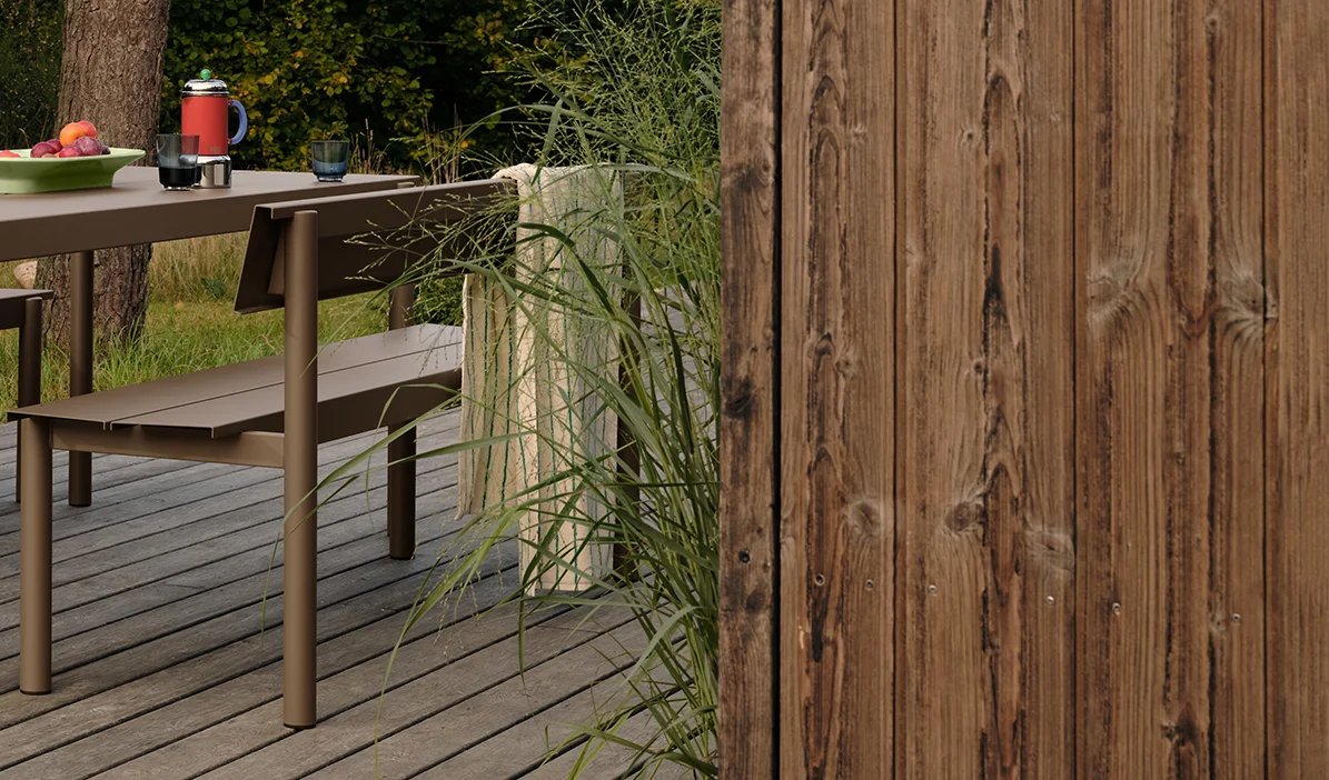 Mesa e banco de metal marrom para exterior em deck de madeira com frutas e bebidas, ao lado de grama alta e cerca de madeira.