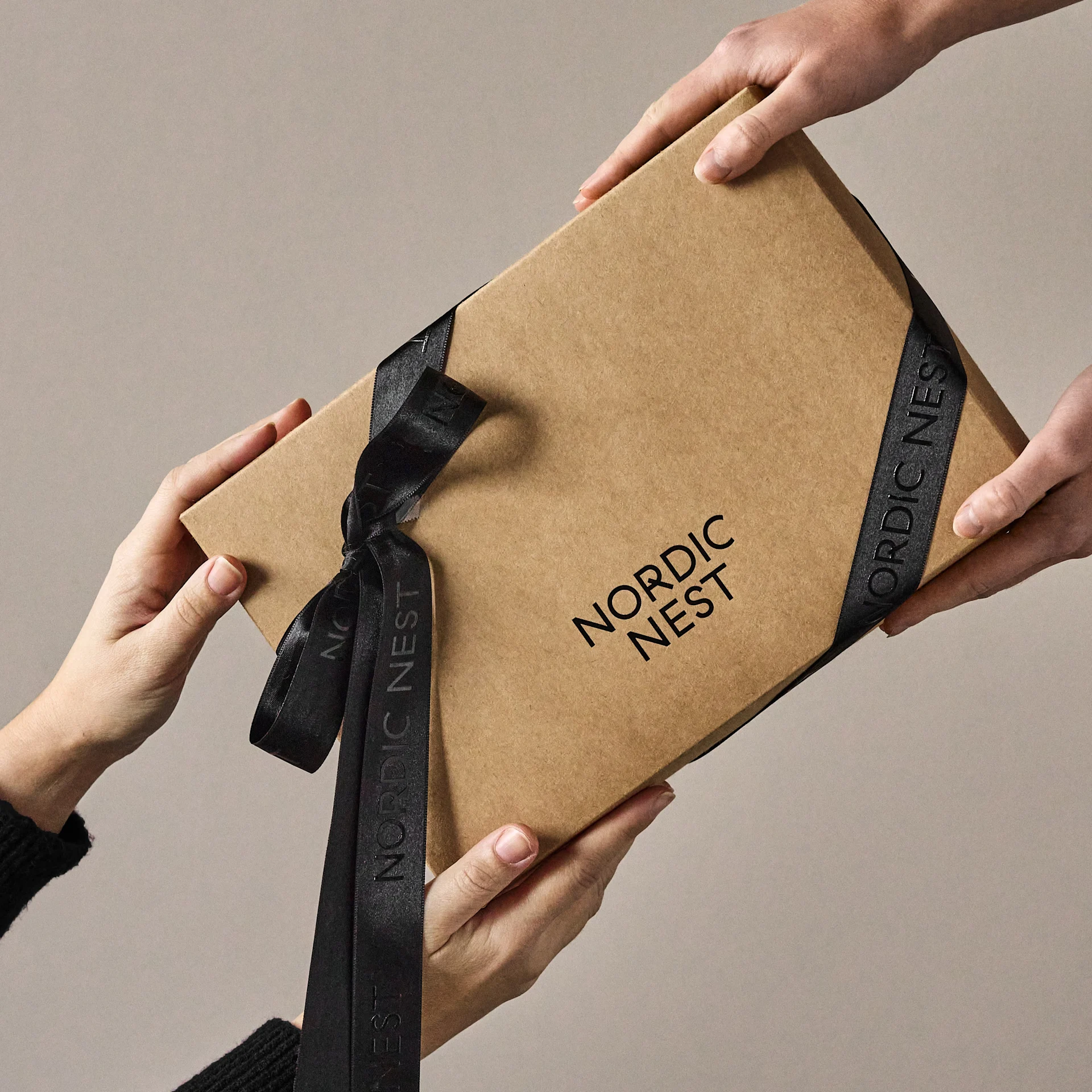 Mãos a trocar uma caixa de presente castanha com o logótipo 'Nordic Nest' e fita preta.