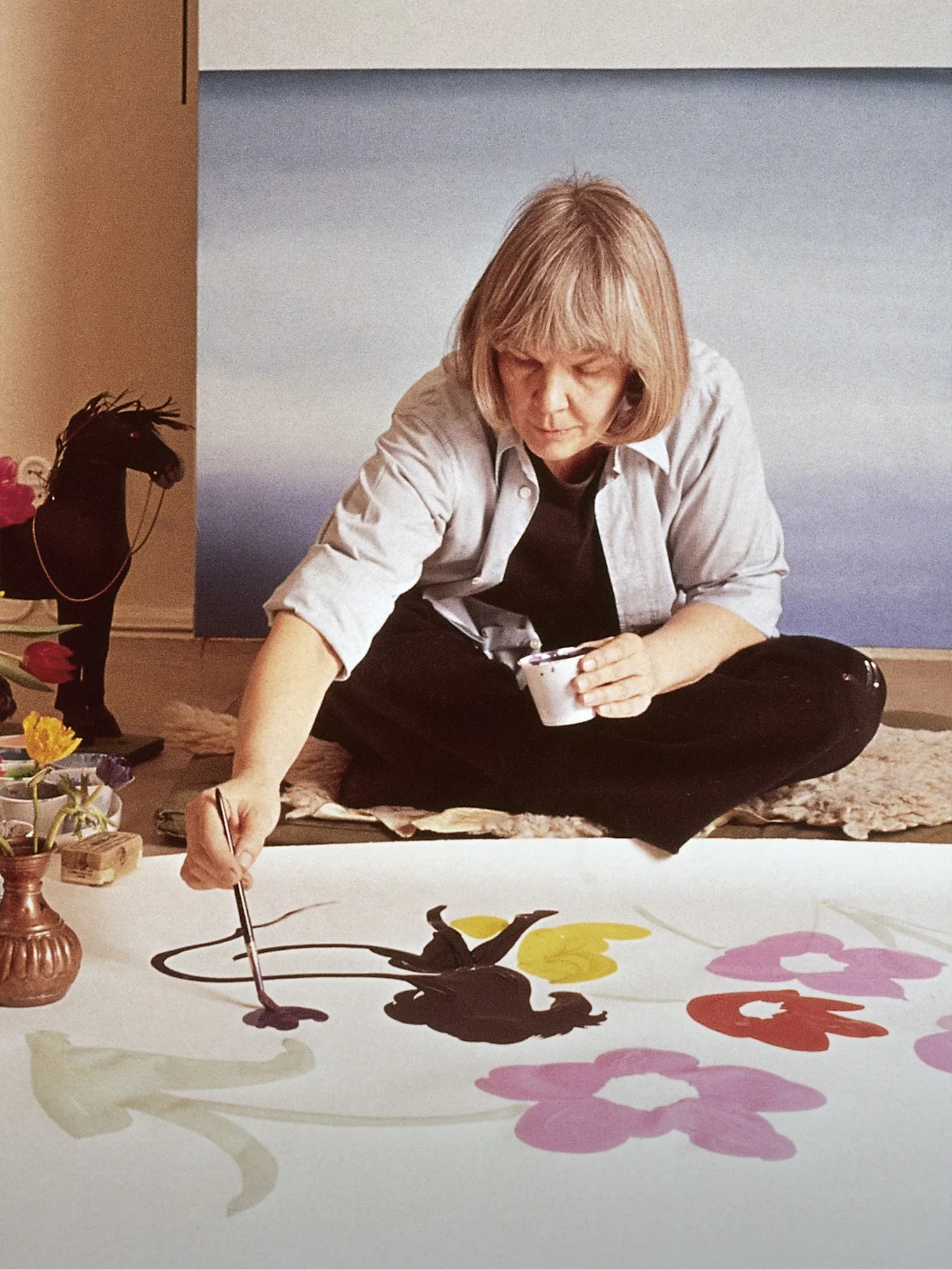 A artista Marimekko Maija Isola sentada no chão pintando grandes flores coloridas em um tecido branco.