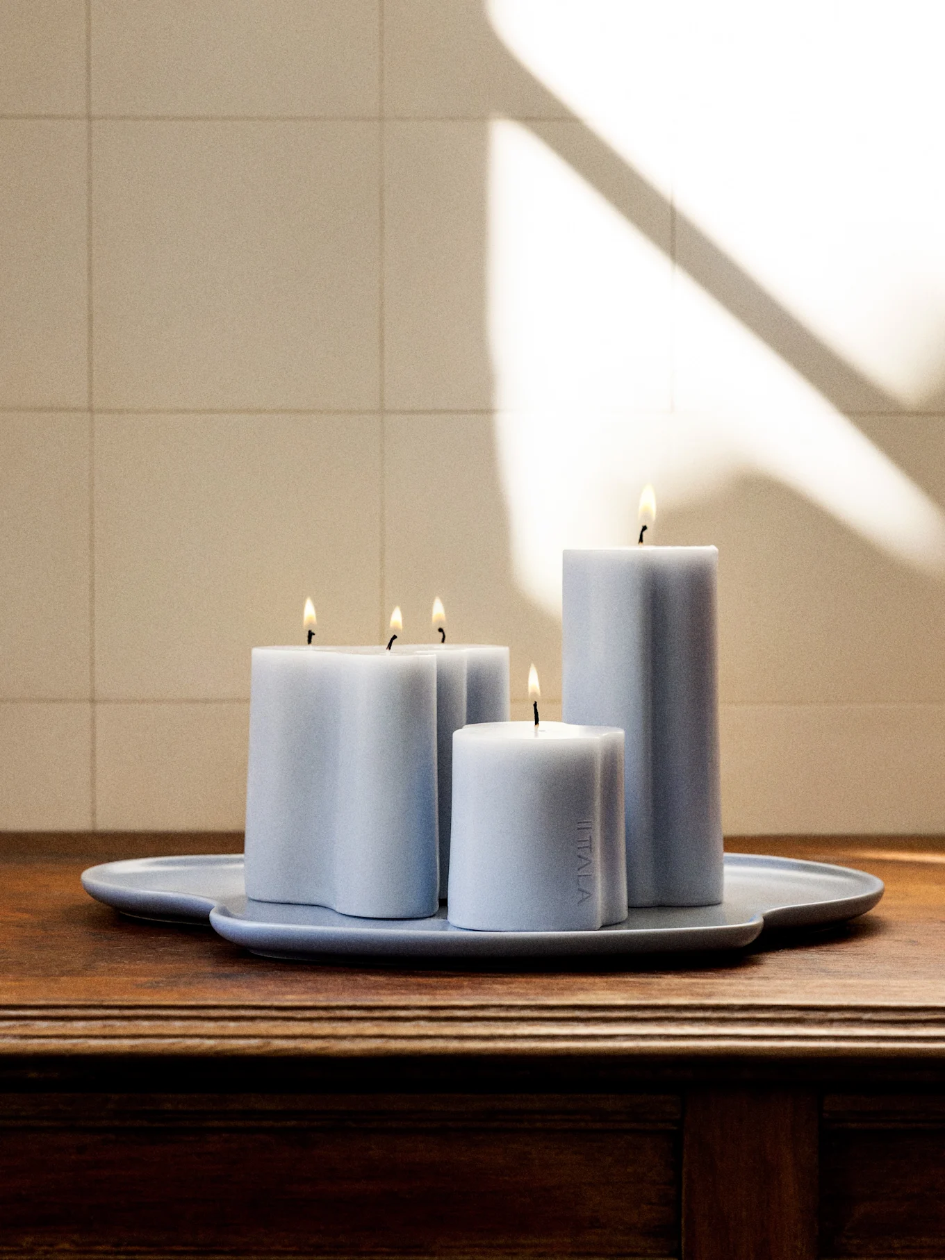 Quatro velas acesas azul-cinzento claro de várias formas e alturas numa bandeja irregular a condizer sobre uma superfície de madeira escura.