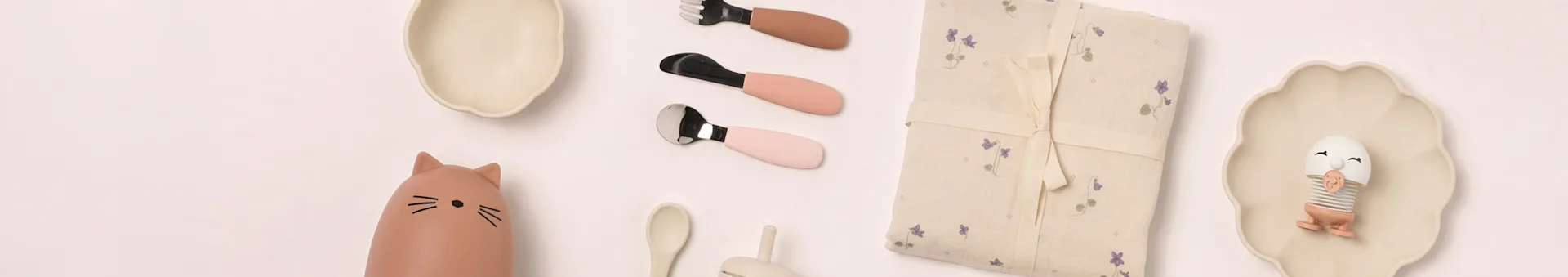 Flat lay de artigos de refeição para bebé: pratos bege, garrafa de gato, talheres rosa, cobertor floral, brinquedo de ovo.