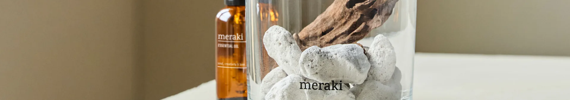 Um frasco marrom de óleo essencial e um vaso transparente com pedras brancas e madeira flutuante. O frasco e o vaso estão ambos rotulados como 'meraki'.